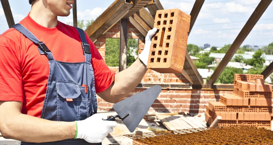 Chantier de construction extérieur avec maçon en salopette bleue et t-shirt rouge tenant une brique perforée et une truelle