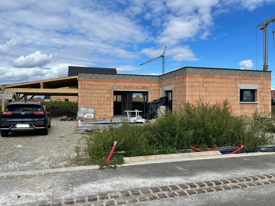 Chantier de construction résidentielle en cours avec maison individuelle en briques terra cotta, toiture partiellement couverte, carport en bois attenant