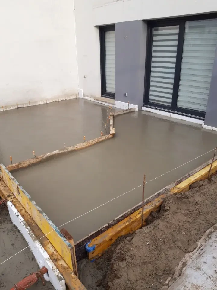 Chantier extérieur de construction d'une terrasse en béton fraîchement coulé devant une façade moderne avec volets roulants gris