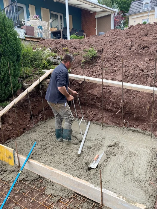 Chantier extérieur de construction avec ouvrier en bottes effectuant le lissage d'une dalle béton fraîchement coulée à l'aide d'une règle de maçon