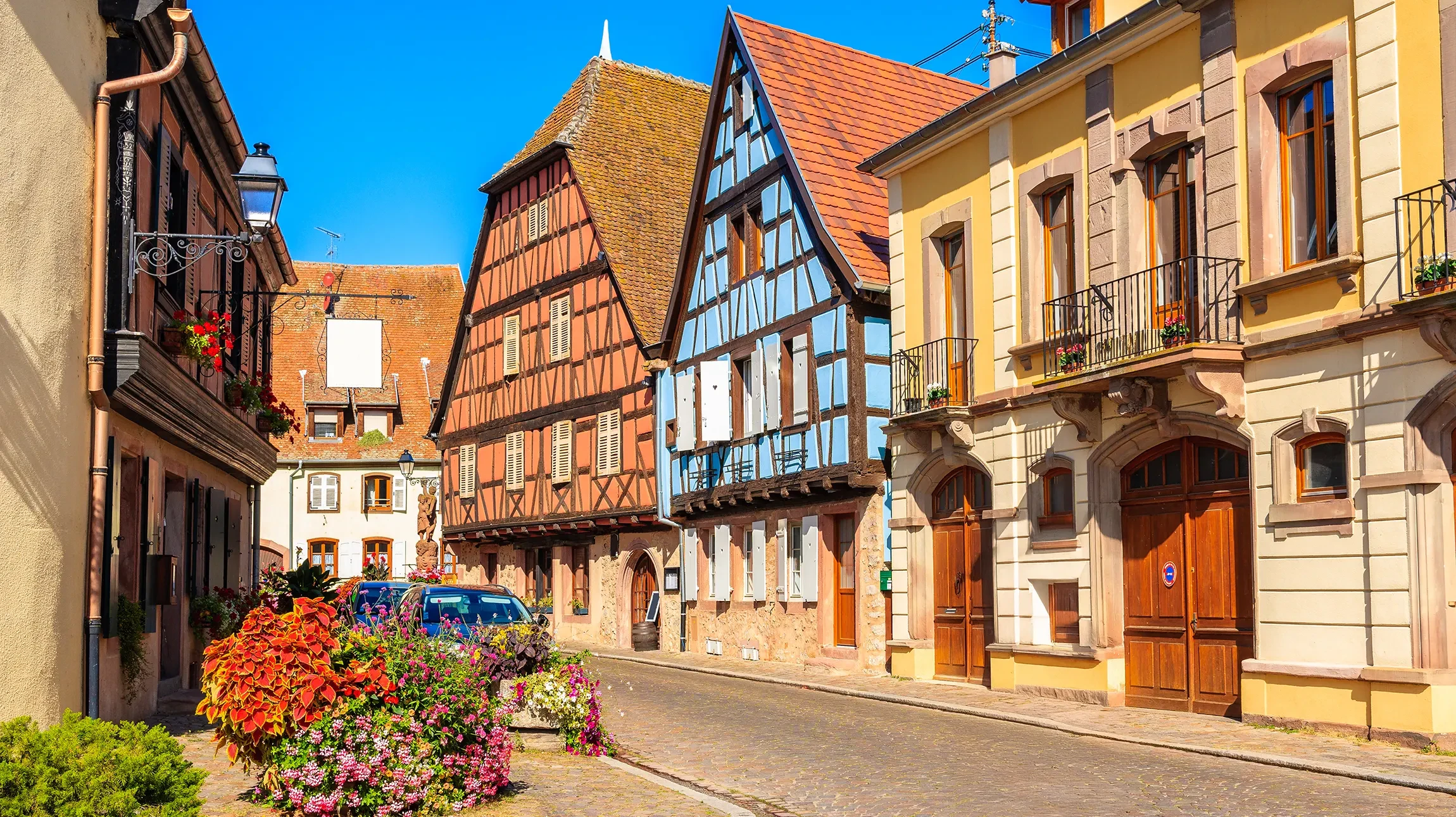Place de village alsacien avec maisons à colombages colorées aux façades bleues, vertes et roses